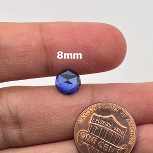 May include: A dark blue round gemstone measuring 8mm in diameter. The gemstone is held in a hand for size comparison next to a UK penny.