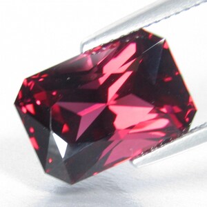 Peut inclure: Une pierre pr&eacute;cieuse rouge fonc&eacute; de forme rectangulaire et &agrave; facettes multiples. La pierre pr&eacute;cieuse est tenue par une pince.