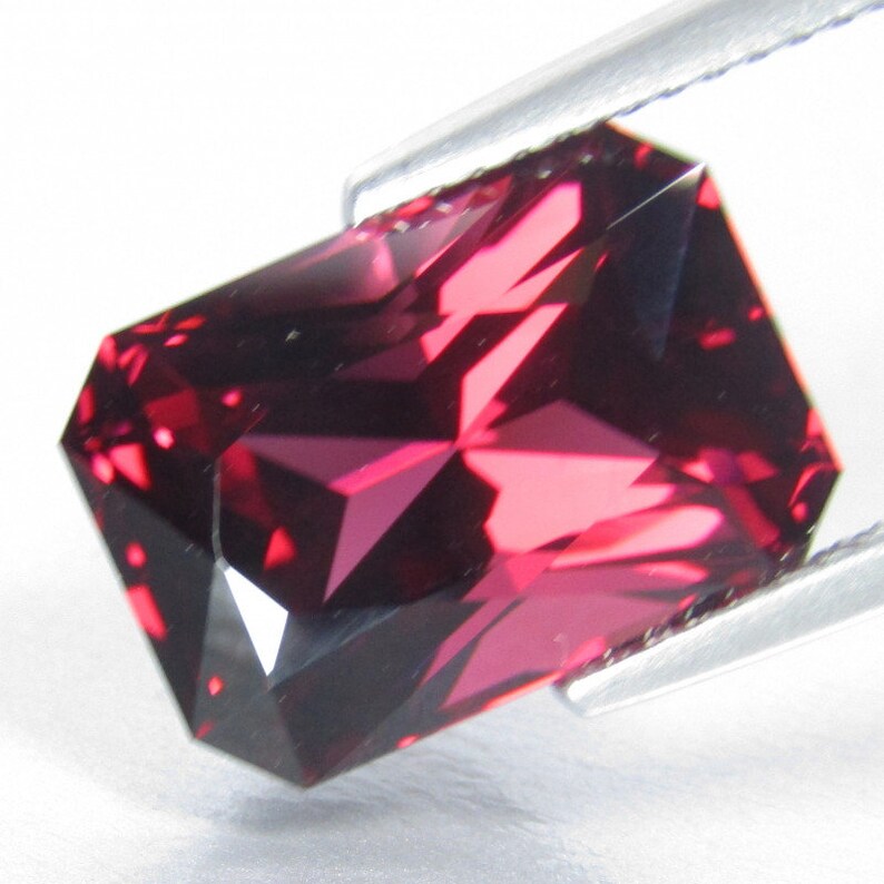 Peut inclure: Une pierre pr&eacute;cieuse rouge fonc&eacute; de forme rectangulaire et &agrave; facettes multiples. La pierre pr&eacute;cieuse est tenue par une pince.