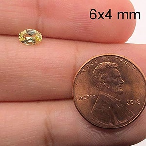 May include: A yellow oval gemstone measuring 6x4 millimetres. The gemstone is being held in a hand for size comparison next to a US penny.