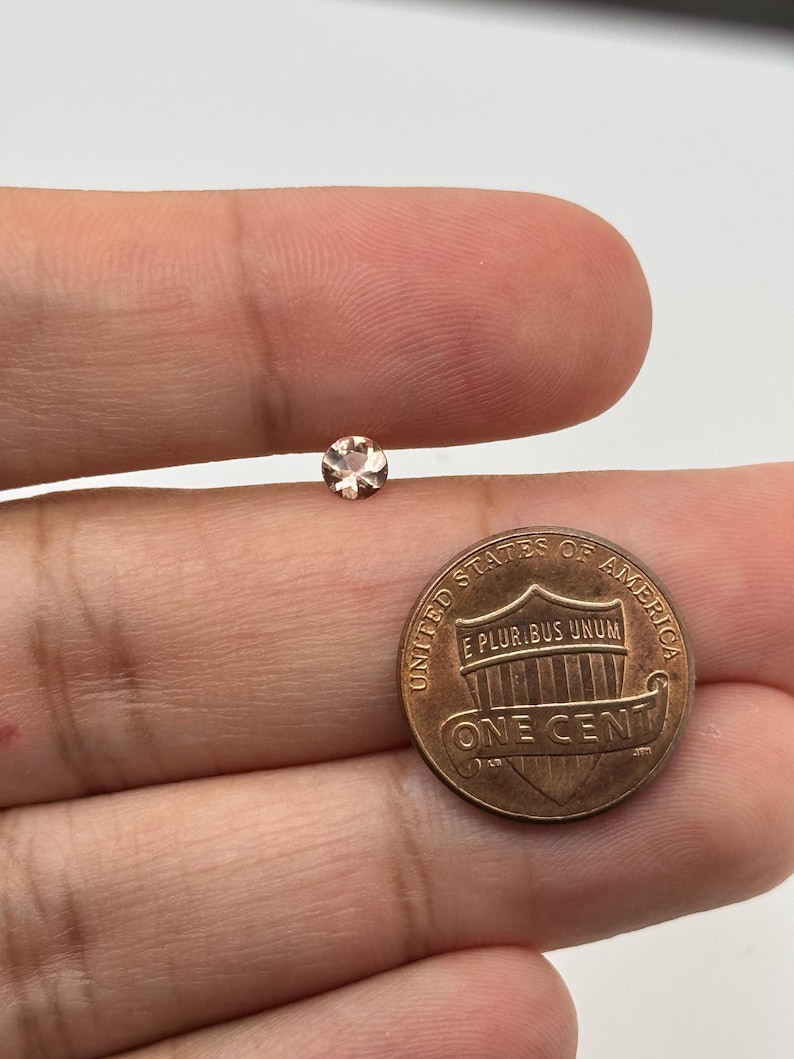Op de afbeelding: Een kleine, ronde, lichtroze edelsteen op een vinger, naast een Amerikaanse cent ter groottevergelijking.