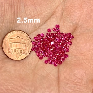 May include: A collection of small, round, bright pink gemstones, approximately 2.5mm in diameter, are displayed next to a United States one-cent coin. The gemstones are scattered across a light-colored surface.