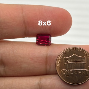 May include: A deep red gemstone with a rectangular shape, measuring 8x6, is held in a hand with a penny for scale.