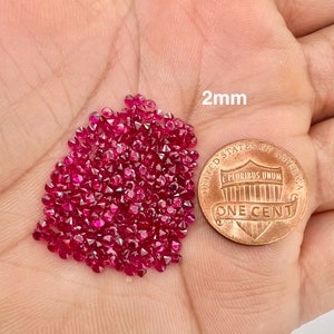 May include: A pile of approximately 100 small, 2mm red ruby gemstones, displayed on a hand for size comparison next to a US penny. The penny has the text "UNITED STATES OF AMERICA" and "E PLURIBUS UNUM" on the top half and "ONE CENT" on the bottom half.