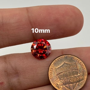 May include: A 10mm round red gemstone with a faceted cut. The gemstone is being held in a hand next to a US penny for size comparison.