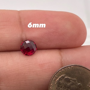 May include: A 6mm round red gemstone with a faceted cut. The gemstone is being held in a hand next to a US penny for size comparison.