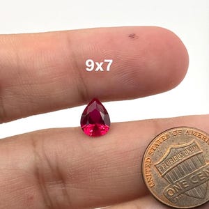 May include: A pear-shaped, faceted, vibrant red gemstone with a 9x7 measurement label, positioned next to a United States one-cent copper coin for scale. The gemstone exhibits a clear, brilliant cut with multiple reflective surfaces.