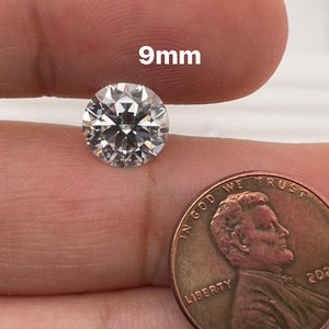 May include: A clear round gemstone measuring 9mm in diameter. The gemstone is being held in a hand next to a US penny for size comparison.