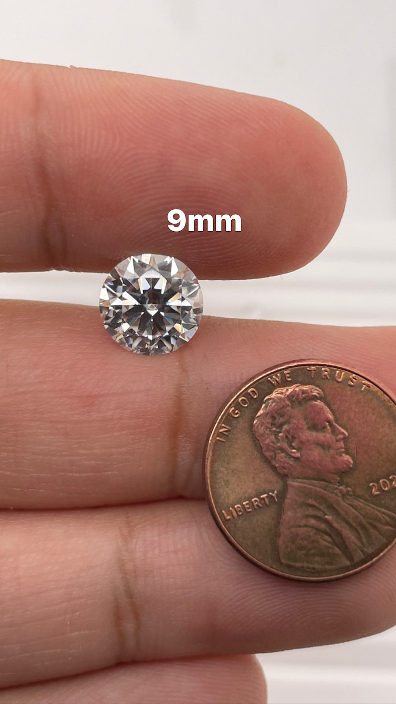 May include: A clear round gemstone measuring 9mm in diameter. The gemstone is being held in a hand next to a US penny for size comparison.