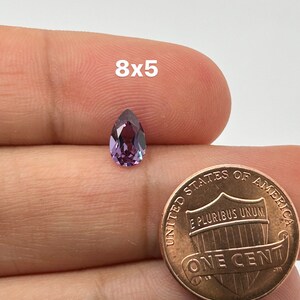 May include: A pear-shaped purple gemstone with a hint of green, measuring 8mm by 5mm, is shown next to a US penny for size comparison.