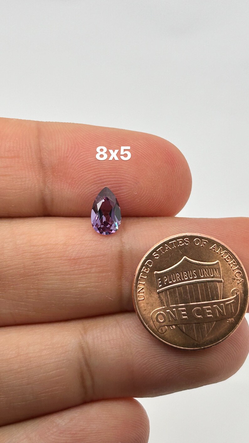 May include: A pear-shaped purple gemstone with a hint of green, measuring 8mm by 5mm, is shown next to a US penny for size comparison.