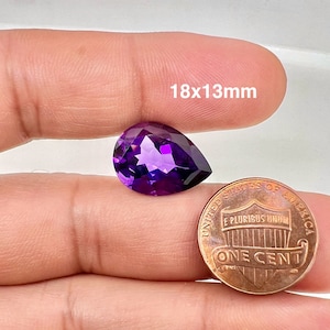 May include: A deep purple pear-shaped gemstone, measuring 18x13mm, is held in a hand next to a US penny for size comparison.