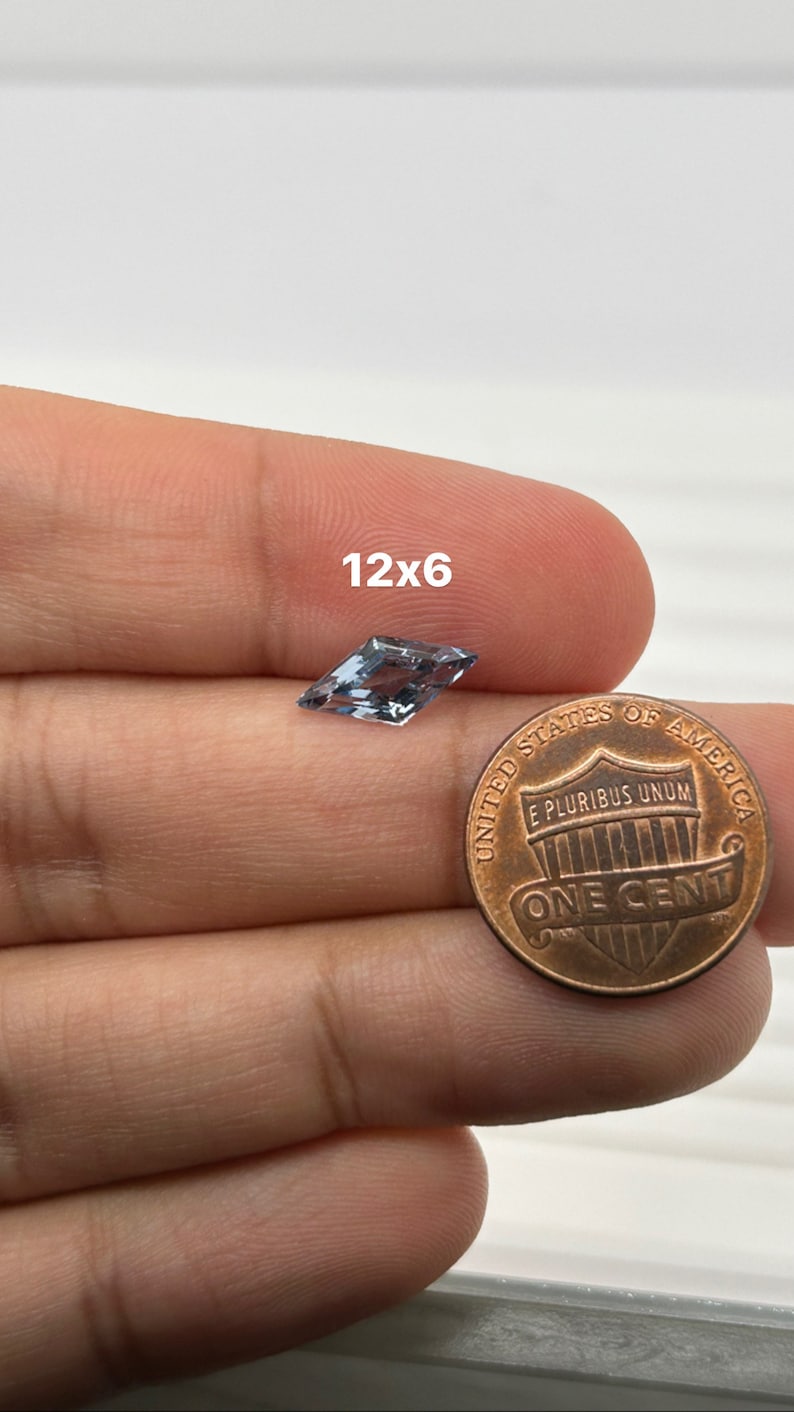 Peut inclure: Une pierre pr&eacute;cieuse bleu clair de forme cerf-volant, mesurant 12x6 mm, est tenue dans une main &agrave; c&ocirc;t&eacute; d'un centime am&eacute;ricain pour comparaison de taille.