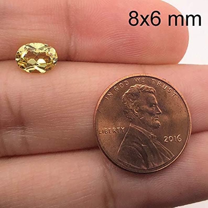 May include: A yellow oval gemstone measuring 8x6 millimetres. The gemstone is held in a hand next to a US penny for size comparison.