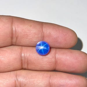 May include: A round, vibrant blue gemstone with a six-rayed star pattern. The star effect is created by light reflecting off inclusions within the stone. The gem is a deep, saturated blue color, and appears to be polished.