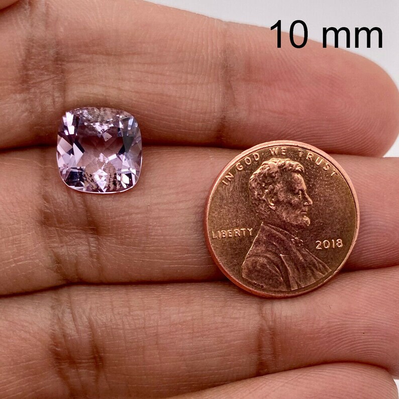 May include: A pale pink cushion-cut gemstone measuring 10 mm in diameter. The gemstone is held in a hand next to a US penny for size comparison.