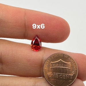 May include: A pear-shaped red gemstone measuring 9x6 mm. The gemstone is held in a hand next to a US penny for size comparison.
