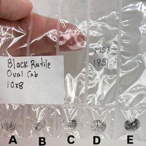 May include: Five oval-shaped black rutile cabochons in individual plastic bags. The bags are labeled with the text "Black Rutile Oval Cab 10x8" and "MS# 1851". The gemstones are labeled A through E. The gemstones have black inclusions.