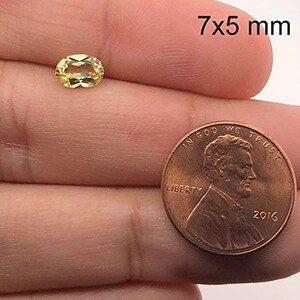 May include: A yellow oval gemstone measuring 7x5 millimetres. The gemstone is held in a hand for size comparison next to a US penny.