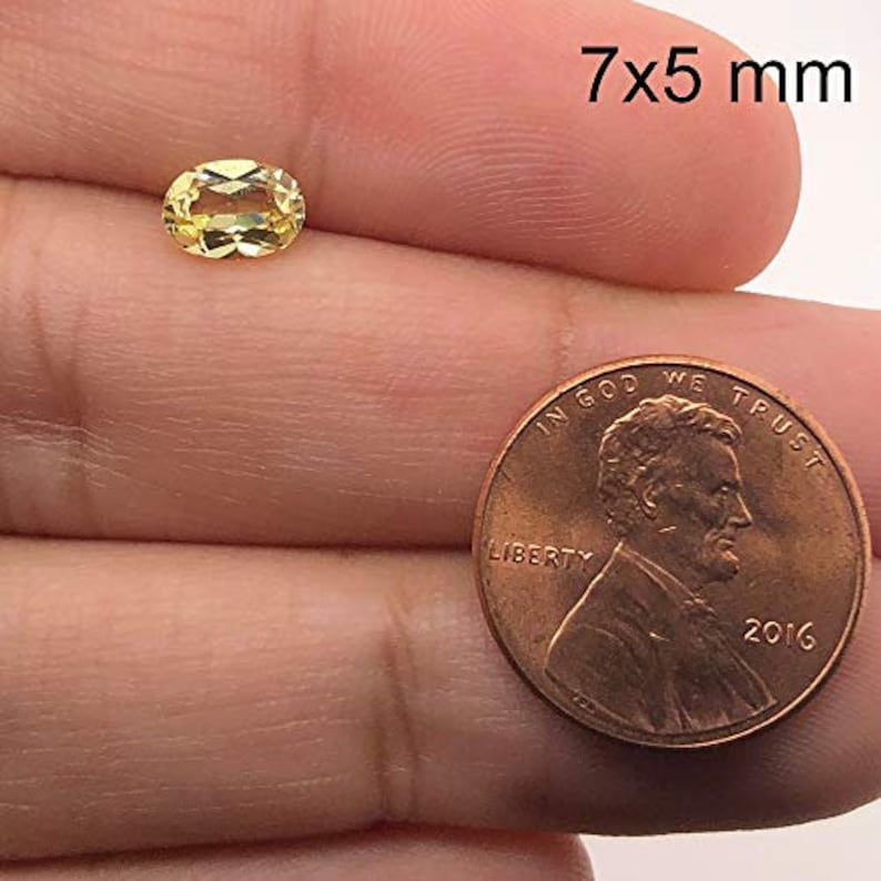 May include: A yellow oval gemstone measuring 7x5 millimetres. The gemstone is held in a hand for size comparison next to a US penny.