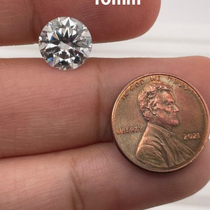 May include: A clear round diamond gemstone measuring 10mm in diameter, next to a US penny for size comparison.
