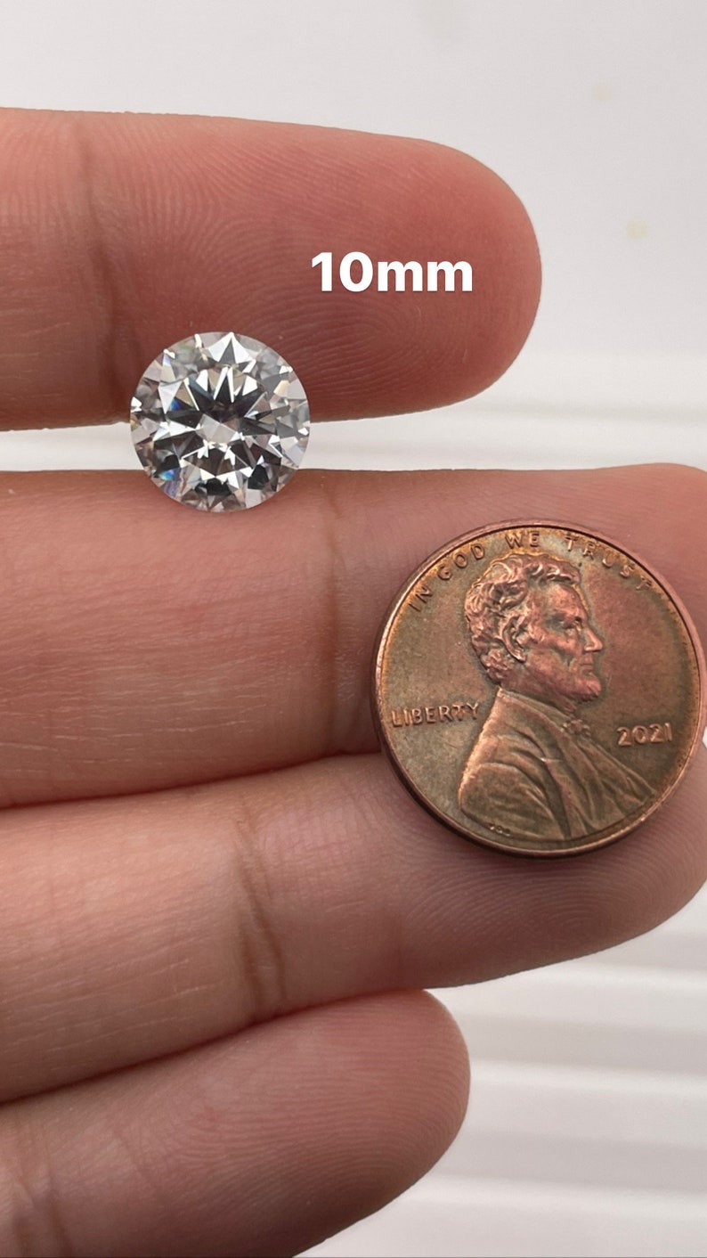 May include: A clear round diamond gemstone measuring 10mm in diameter, next to a US penny for size comparison.