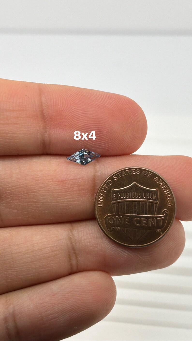 Peut inclure: Une pierre pr&eacute;cieuse bleu clair de forme rectangulaire, mesurant 8x4 millim&egrave;tres, est tenue dans une main &agrave; c&ocirc;t&eacute; d'un penny am&eacute;ricain pour comparaison de taille.