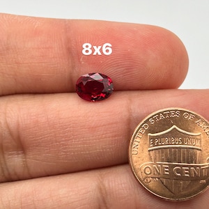 May include: A deep red oval gemstone, measuring 8x6 millimeters, is held in a hand next to a US penny for size comparison.