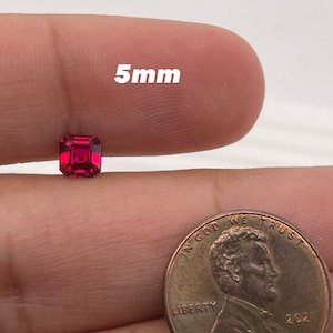 May include: A 5mm square red gemstone, held in a hand next to a US penny for size comparison.