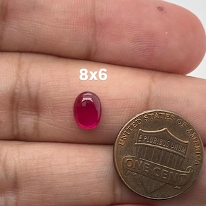 May include: A small, oval-shaped, deep pink gemstone with a smooth, polished cabochon surface. The stone is positioned next to a United States one-cent coin for scale, with the text 8x6 printed clearly above the gemstone to indicate its size.
