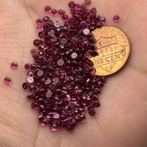 May include: A collection of small, faceted, deep red gemstones, possibly garnets, are scattered across a surface. A copper one-cent coin is partially visible, providing a size reference. The gems have a brilliant, polished appearance.