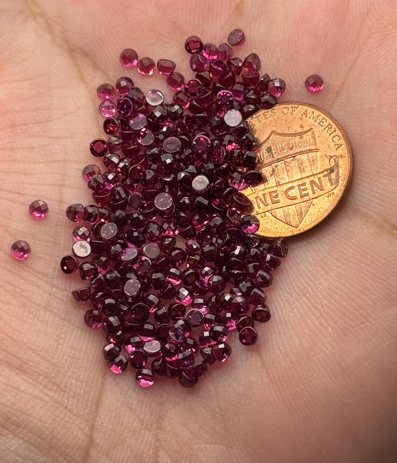 May include: A collection of small, faceted, deep red gemstones, possibly garnets, are scattered across a surface. A copper one-cent coin is partially visible, providing a size reference. The gems have a brilliant, polished appearance.