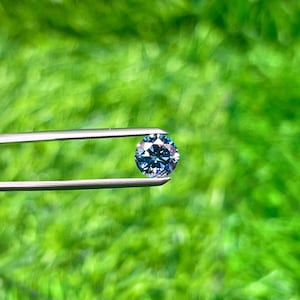 Puede incluir: Una gema azul redonda y facetada, sostenida por unas pinzas plateadas sobre un fondo verde borroso. La gema tiene múltiples facetas que reflejan la luz, creando un efecto brillante. Las pinzas están enfocadas, resaltando los detalles de la gema.