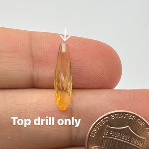 May include: A close-up of a faceted, orange gemstone with a drill hole at the top. The gemstone is being held by a hand, and a US penny is in the background for size comparison. Top drill only.