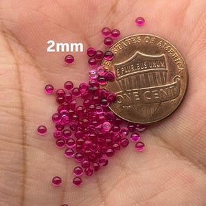 May include: A pile of approximately 50 small, round, 2mm ruby red beads next to a US penny. The beads are shiny and have a smooth surface.