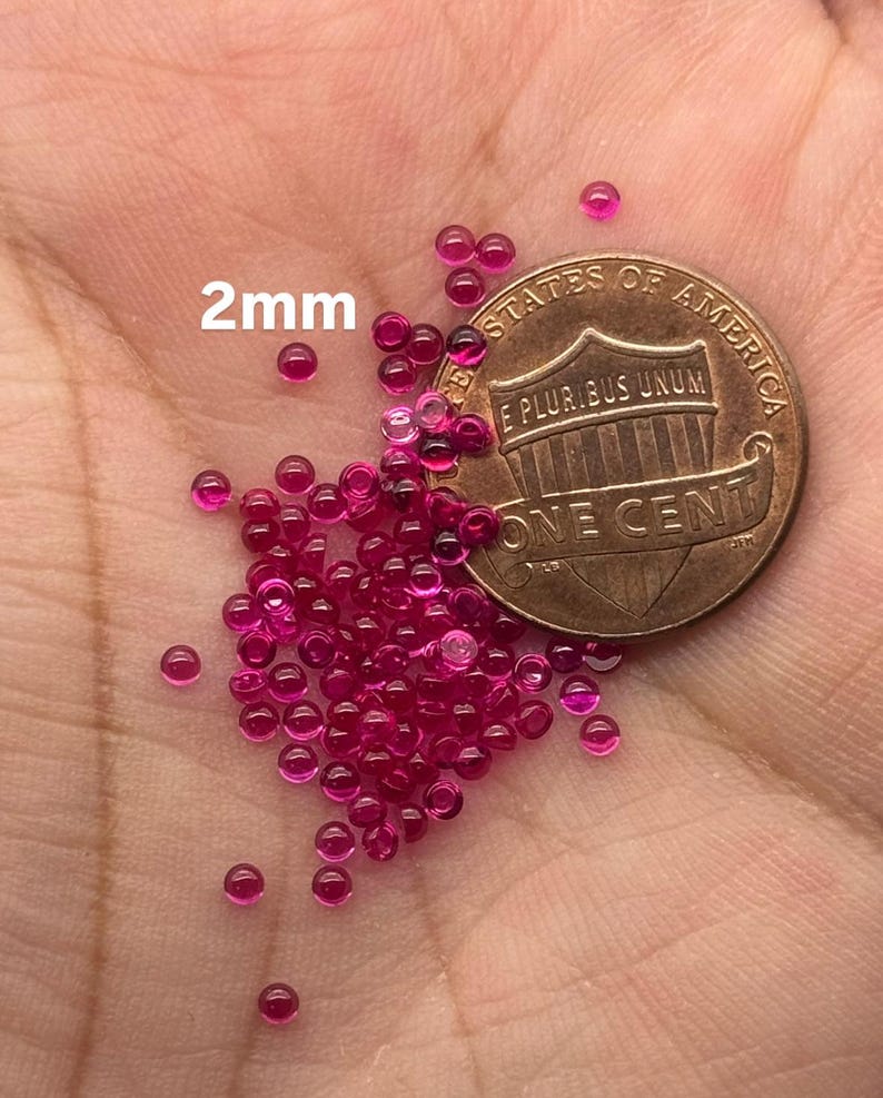 May include: A pile of approximately 50 small, round, 2mm ruby red beads next to a US penny. The beads are shiny and have a smooth surface.