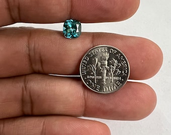May include: A teal blue gemstone with a square cushion cut, held in a hand next to a US dime for size comparison.