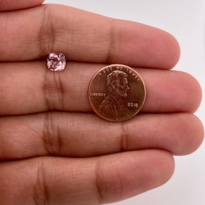 May include: A pink gemstone with a cushion cut, next to a US penny with the inscription "In God We Trust" and the year "2018".