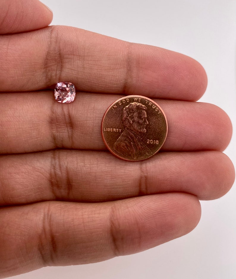 May include: A pink gemstone with a cushion cut, next to a US penny with the inscription "In God We Trust" and the year "2018".