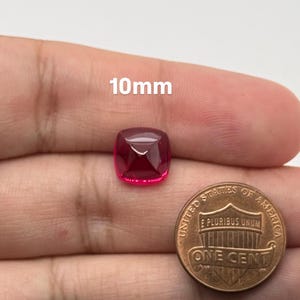 May include: A cushion-cut, deep pink gemstone measuring 10mm in size, positioned next to a United States one-cent copper coin for scale. The gemstone features a faceted surface that reflects light, showcasing a vibrant, saturated color.