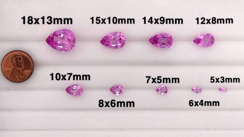 May include: A collection of pink pear-shaped gemstones of various sizes, ranging from 5x3mm to 18x13mm. The gemstones are arranged in rows on a white surface, with a penny for scale.
