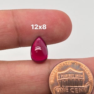 May include: A deep red teardrop-shaped cabochon gemstone measuring 12x8mm. The gemstone is held in a hand for size comparison next to a US penny.