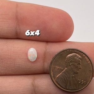 May include: A white oval opal gemstone measuring 6x4 millimeters, held in a hand next to a 1990 US penny for size comparison.