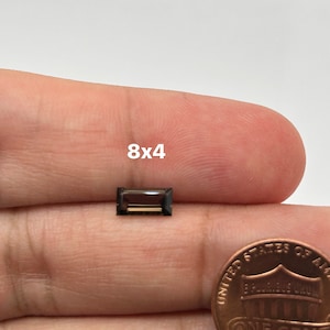 Puede incluir: Una piedra preciosa rectangular de color marrón oscuro con un centro marrón ligeramente más claro, que mide 8x4 mm, sostenida en una mano junto a un centavo estadounidense.