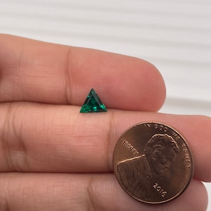 May include: A green triangular gemstone held in a hand next to a US penny.
