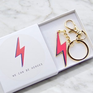 May include: A white gift box with a pink and blue lightning bolt design and the text "We can be heroes". The box contains a gold key chain with a pink lightning bolt charm.