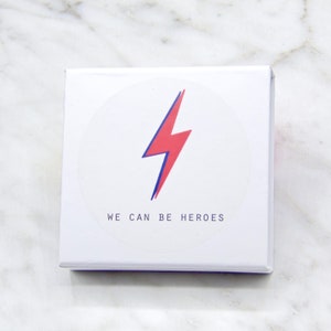 May include: White box with a red lightning bolt graphic and the text "WE CAN BE HEROES" in a circle.