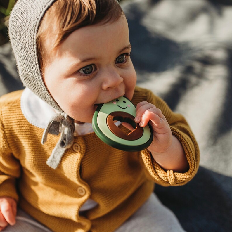 Puede incluir: Un beb&eacute; con un gorro de punto gris y un c&aacute;rdigan amarillo est&aacute; mordiendo un mordedor en forma de aguacate verde con una semilla marr&oacute;n y una cara sonriente.