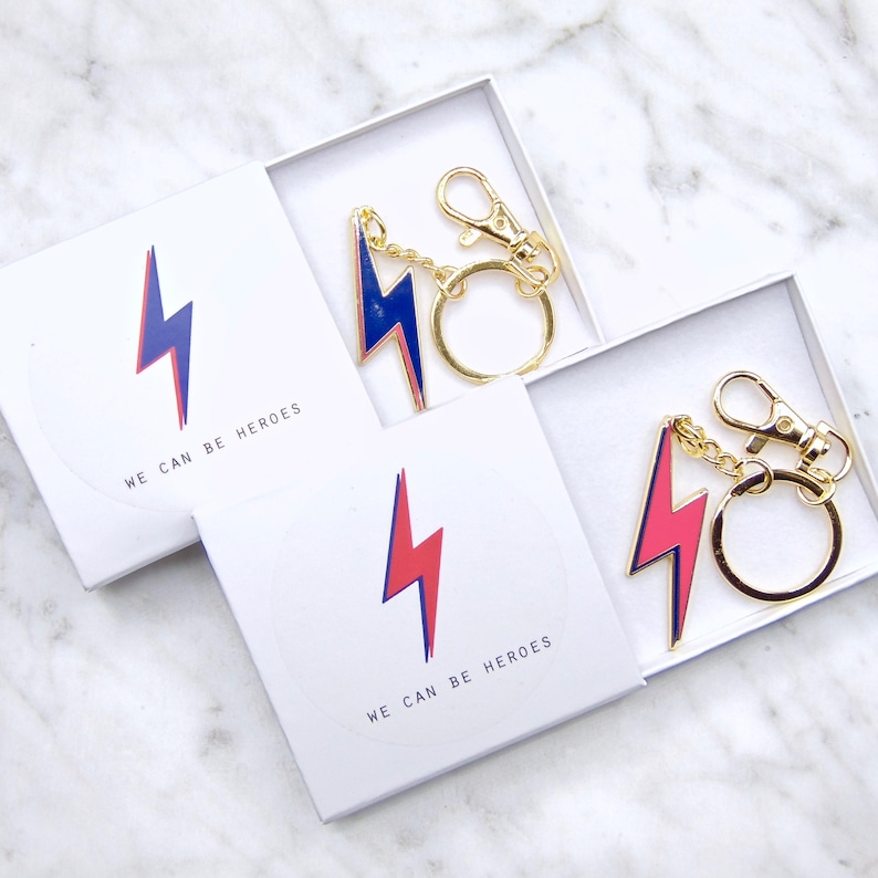 May include: Two white boxes with gold keychains featuring a blue and red lightning bolt design. The boxes have the text "We can be heroes" printed on them.