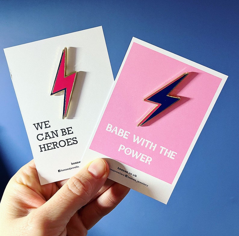 May include: Two enamel pins with lightning bolt designs. The pin on the left is pink with gold trim and says "We can be heroes". The pin on the right is blue with gold trim and says "Babe with the power".
